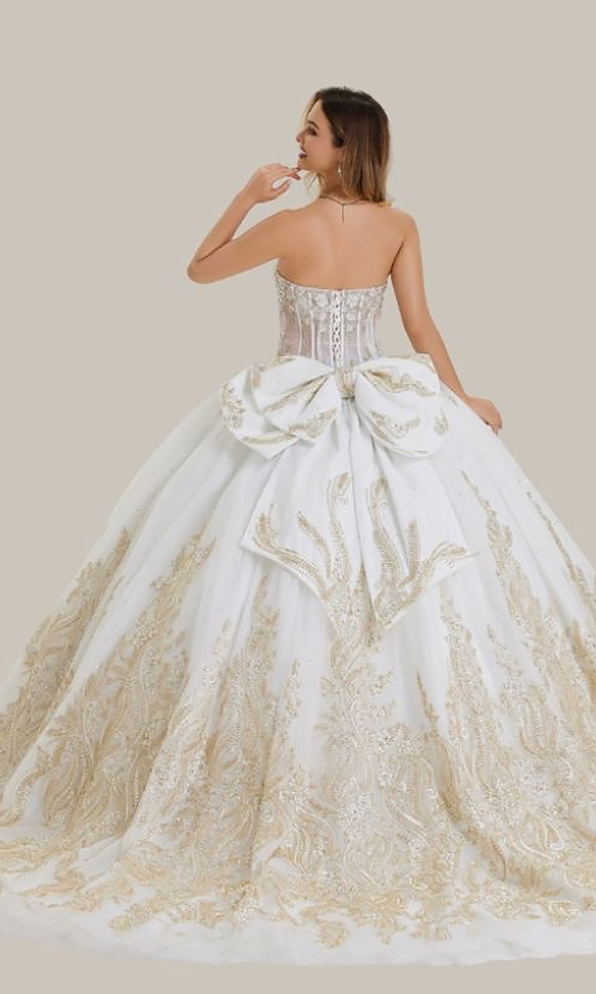 A woman wearing the Quinceanera Dress 26405 by Stella Couture, a strapless white ball gown with a corset bodice, gold embellishments, and a large back bow, stands against a plain beige background.